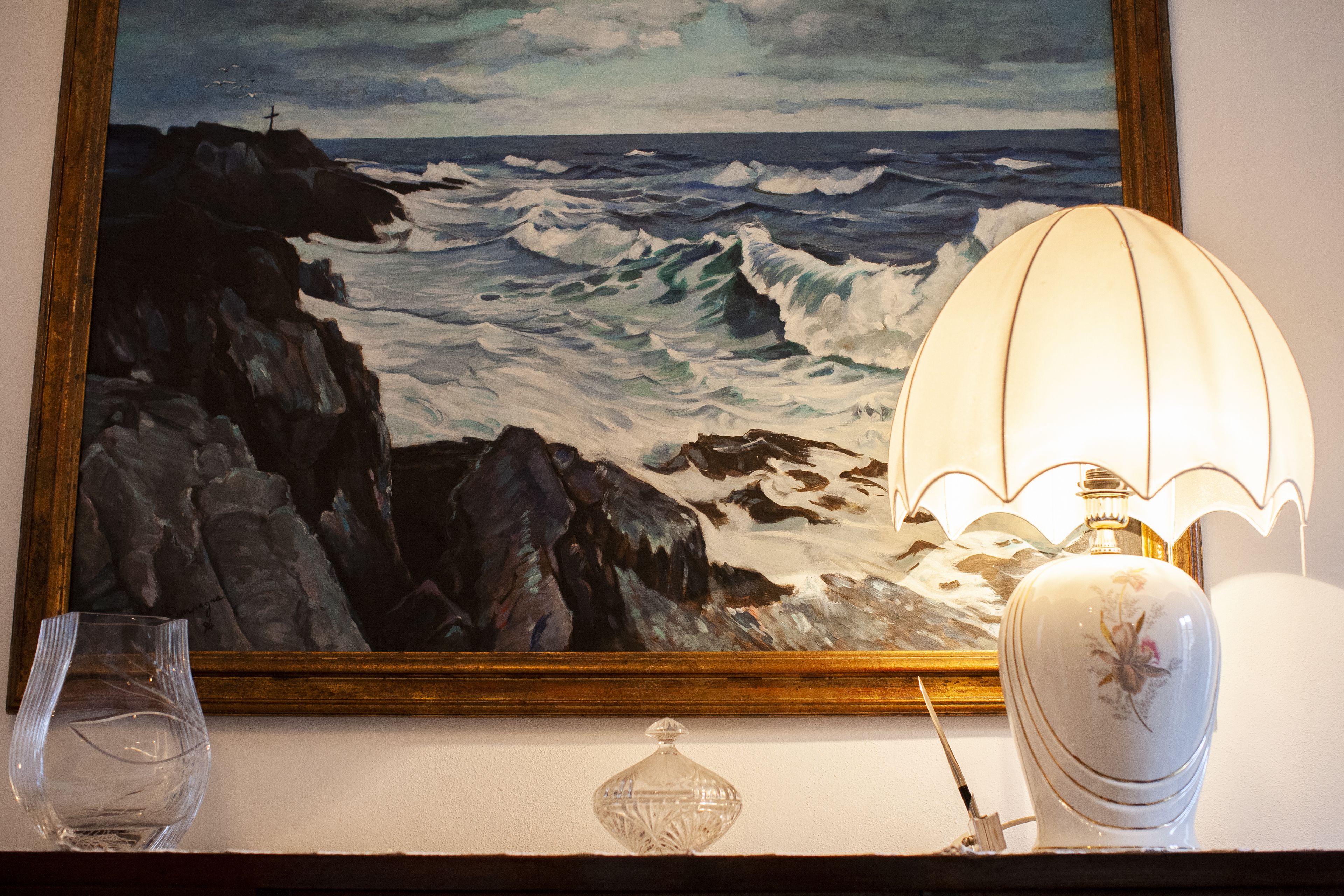 A framed painting of a coastal scene with waves, a lighthouse, and a cross on a rock.