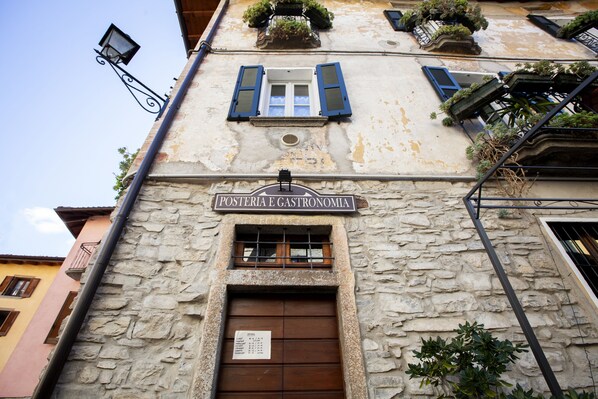 Un bâtiment à la façade en pierre, une enseigne indiquant « POSTERIA E GASTRONOMIA », et une fenêtre protégée par une grille métallique.