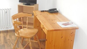 Ein kleiner, ordentlicher Raum mit einem Holztisch, einem Stuhl, einem Fernseher auf einem Standfuß und einer Heizung.