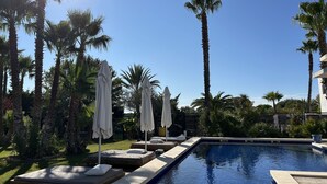 Un espace piscine agrémenté de chaises longues et de parasols, entouré de palmiers et surplombé d’un ciel dégagé.