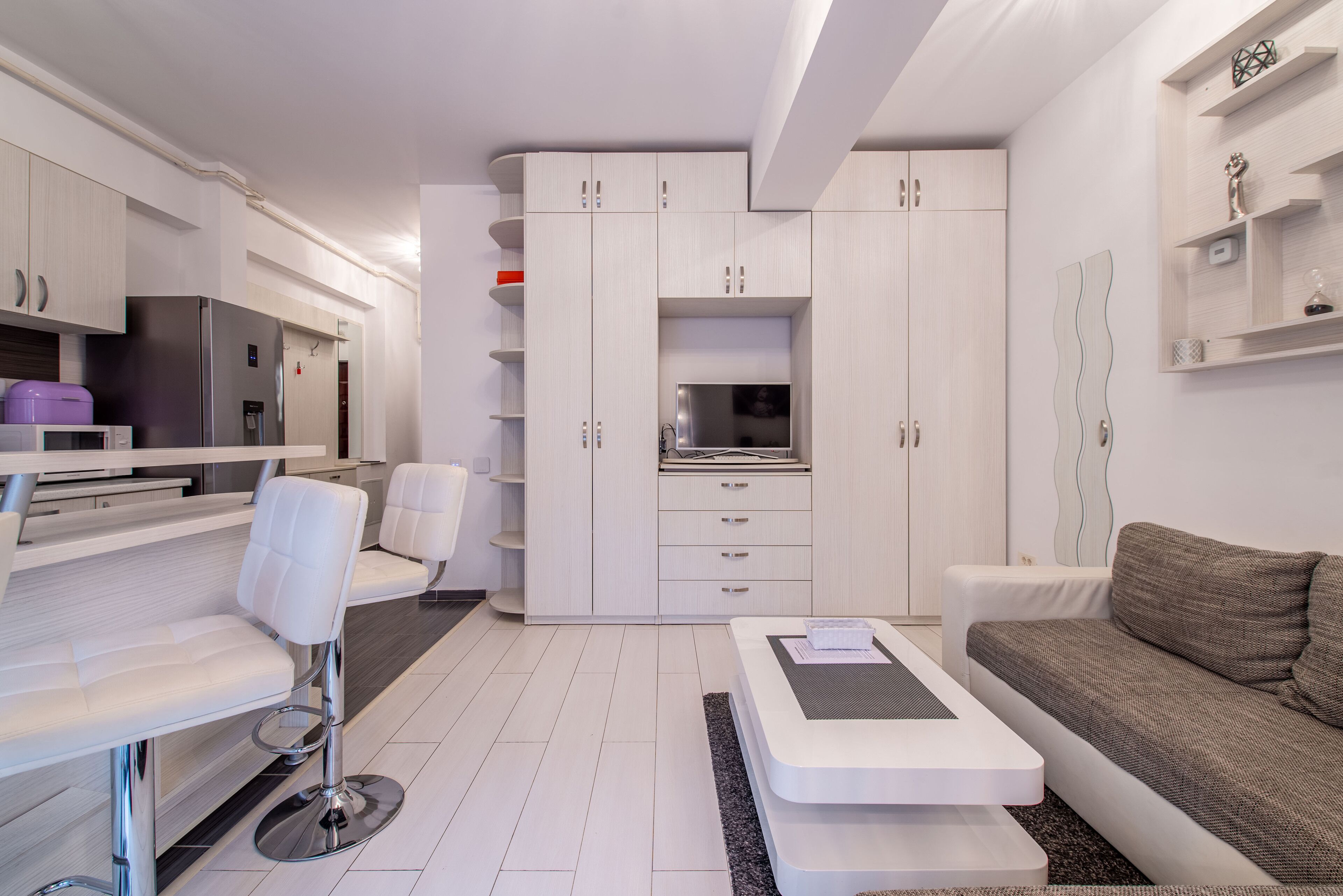 A modern kitchen with white cabinetry, a built-in microwave, and a refrigerator. There is a dining area with a white table and chairs, and a gray sofa in the living area.