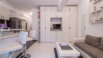 A modern kitchen with white cabinetry, a built-in microwave, and a refrigerator. There is a dining area with a white table and chairs, and a gray sofa in the living area.
