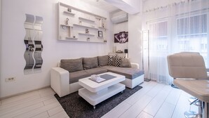 Una sala de estar moderna con un sofá seccional gris, una mesa de centro blanca y un sillón blanco. Hay un estante montado en la pared con objetos decorativos y un espejo grande.