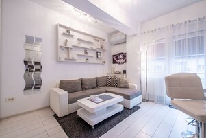 Una sala de estar moderna con un sofá seccional gris, una mesa de centro blanca y un sillón blanco. Hay un estante montado en la pared con objetos decorativos y un espejo grande.