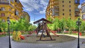 Un parque infantil con columpios y toboganes, rodeado de una zona pavimentada con bancos y farolas, con edificios residenciales al fondo.