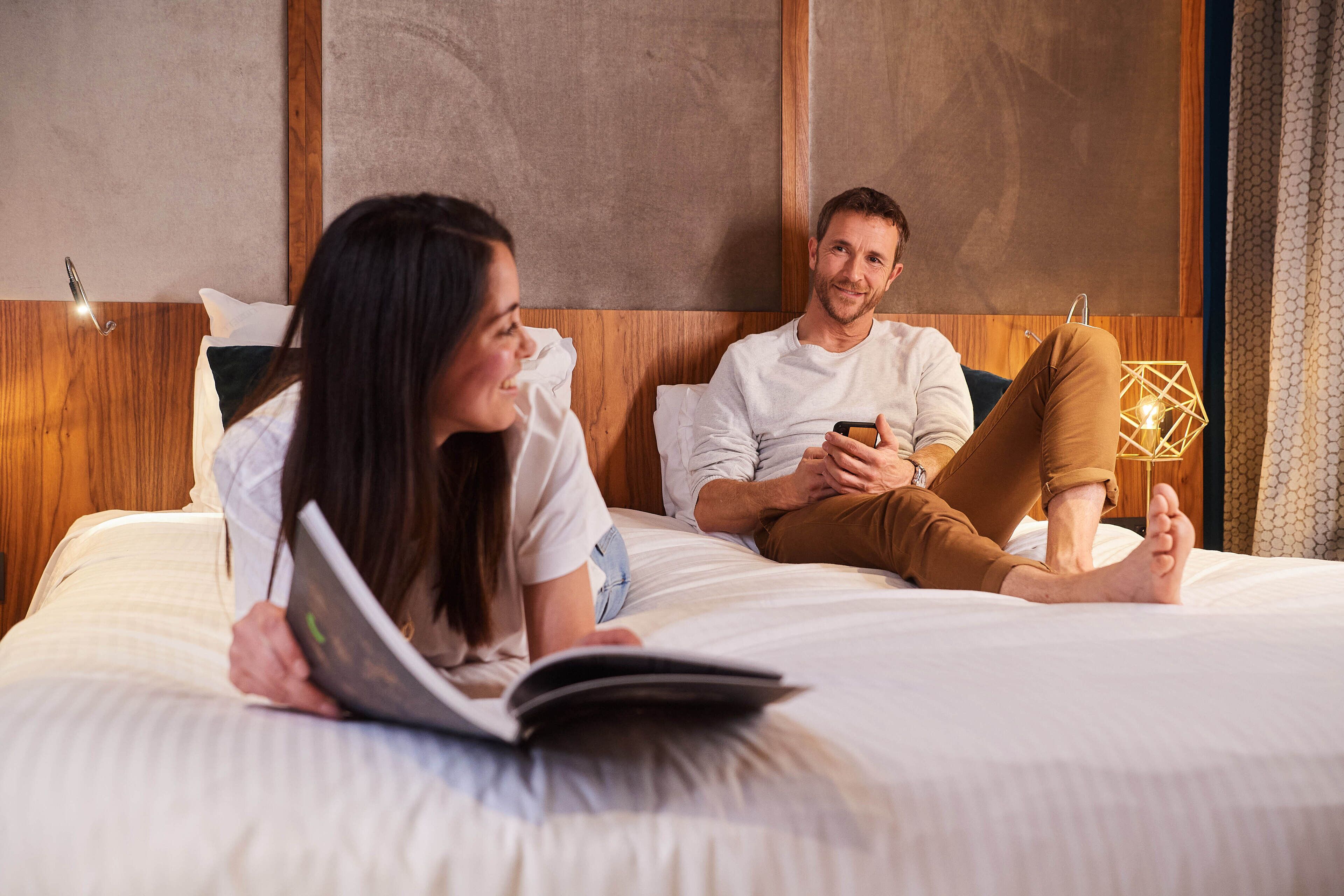 A couple lying on a bed in a hotel room, reading a magazine.