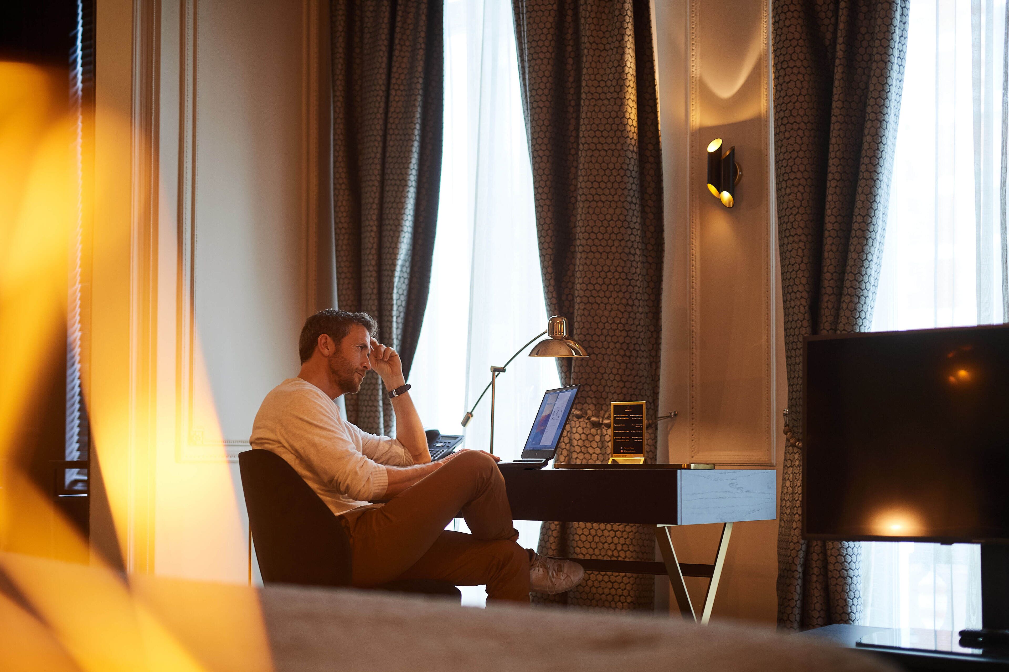 Een man zit op een bed in een hotelkamer, en gebruikt een laptop en een smartphone.