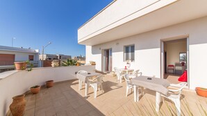 Una terraza en la azotea con mobiliario blanco, plantas en macetas y un cielo azul despejado.