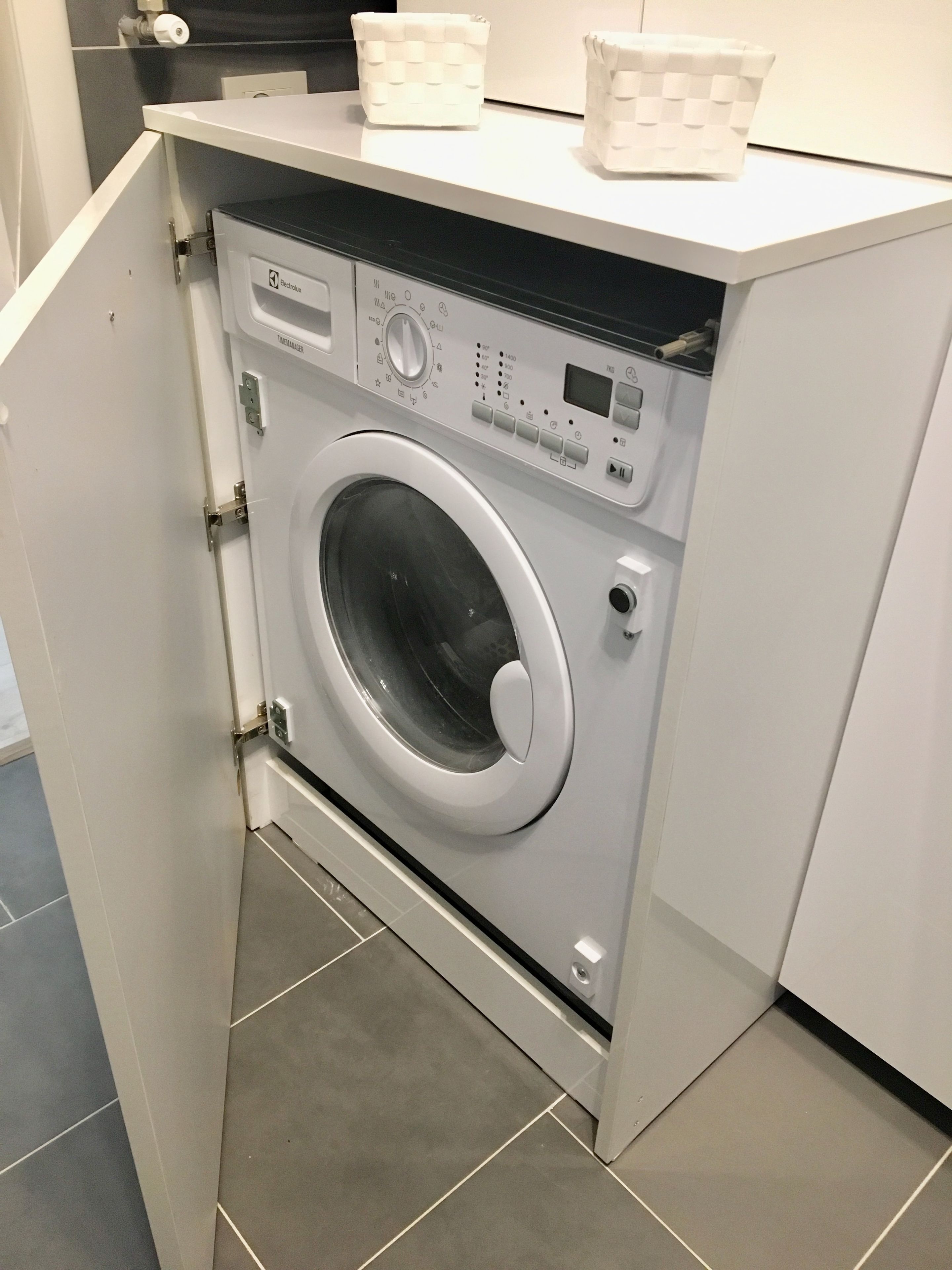 A compact, modern laundry room with a front-loading washing machine, a control panel, and a small storage area above the machine.