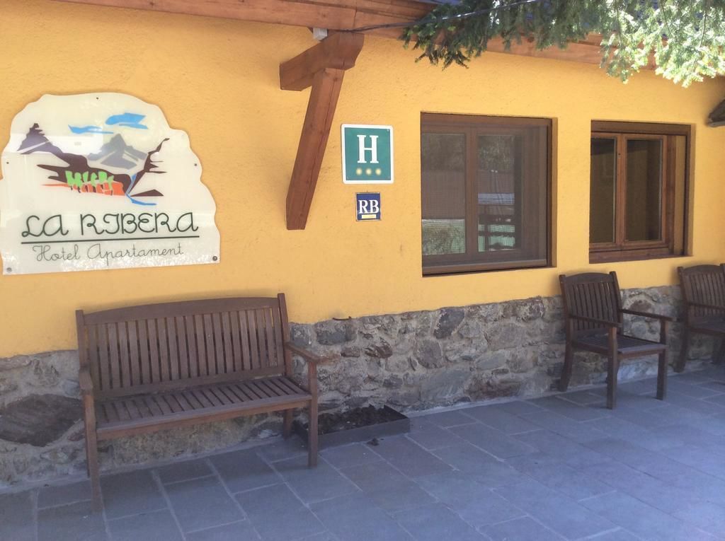 Edifici groc amb un cartell de l'Apart Hotel La Riberas, una banca de fusta i dues cadires.