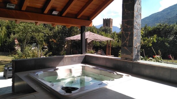 Un bain à remous sous une pergola en bois, soutenue par un pilier en pierre, entouré de verdure et offrant une vue sur la montagne.
