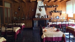 Un espace repas aux murs et au plafond en bois, avec des tables recouvertes de nappes et des chaises, ainsi qu’une cheminée surmontée d’un luminaire suspendu.