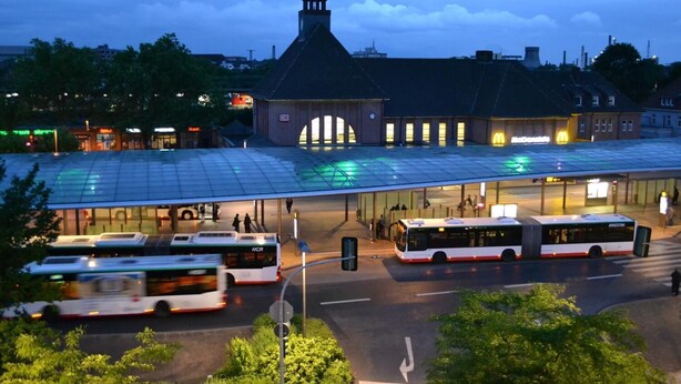 Ein Busbahnhof mit mehreren Bussen, einem überdachten Wartebereich und einem Gebäude mit einem Turm.