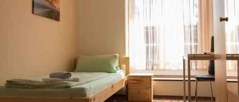 Une petite chambre avec un lit simple, un plancher en bois, un bureau et une chaise.