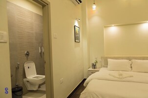 A hotel bathroom with a toilet, shower, and a bed with white linens.