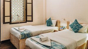 Ein Zimmer mit zwei Einzelbetten, jeweils ausgestattet mit einem Handtuch aufgerollt und einem gemusterten Kopfkissen. Ein kleiner Tisch mit Snacks und einer Lampe ist zu sehen.