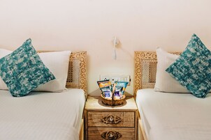Ein Hotelzimmer mit zwei Betten, jedes mit einem weißen Kopfkissen und einem Kissen in einem blaugrünen Muster. Ein Holznachttisch mit einem Korb, der Snacks und eine Flasche enthält.