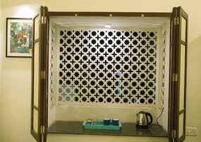 A room with a window featuring a geometric pattern, a small table with a tray holding two cups and a kettle, and a framed picture on the wall.