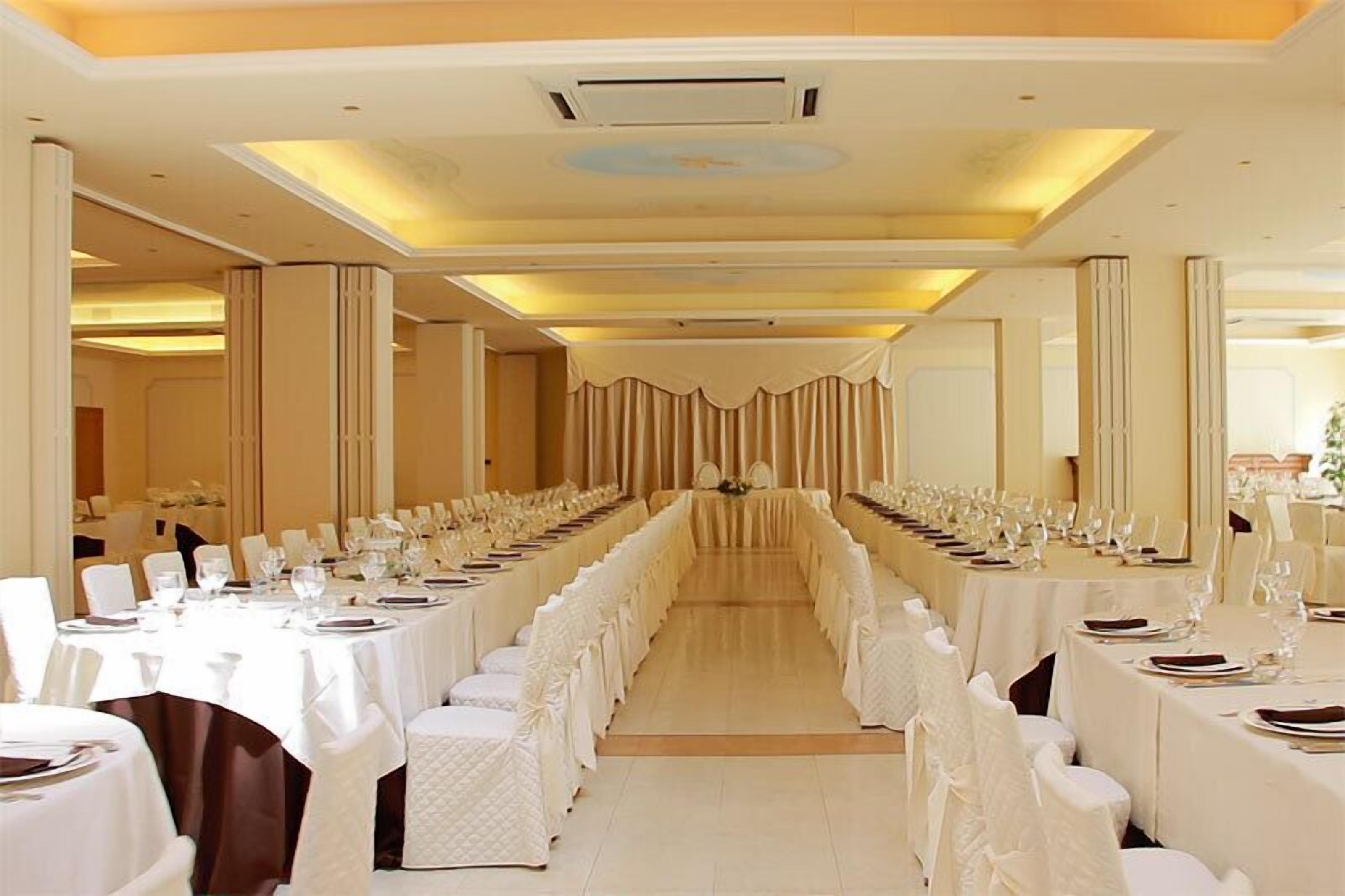 Une longue salle de banquet où les tables sont disposées pour un événement formel, avec une allée centrale et des colonnes disposées de manière symétrique.