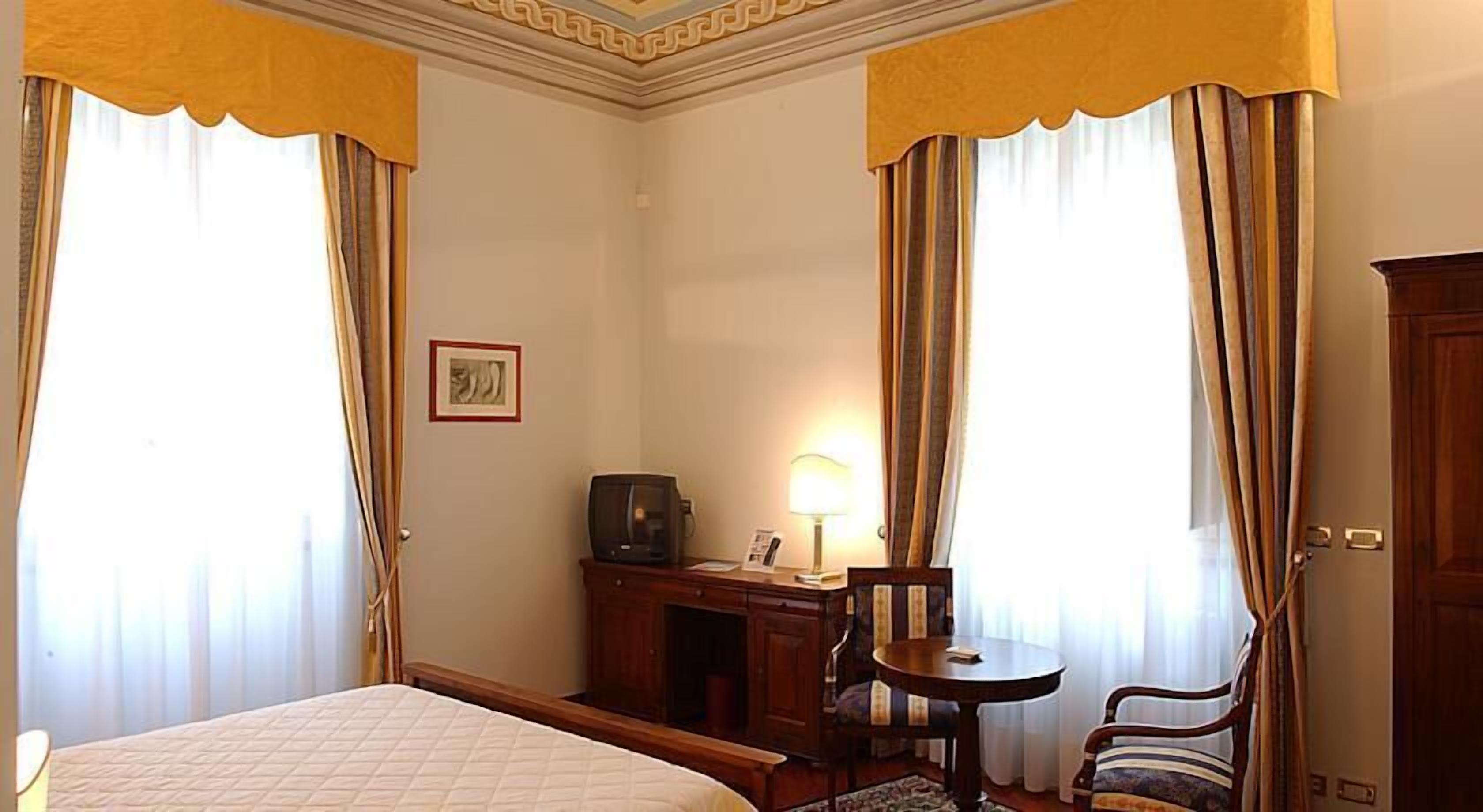 A hotel room with a bed, a desk with a lamp, a chair, and curtains.