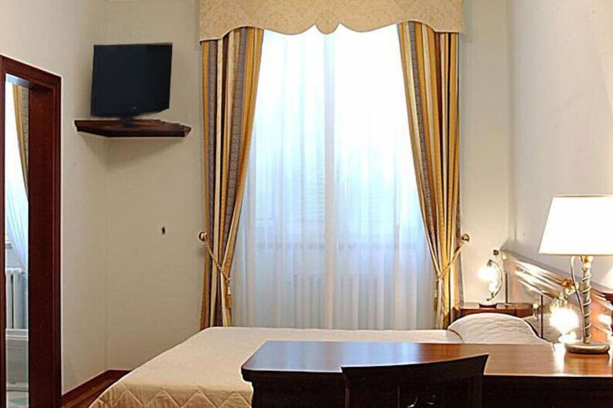 A hotel room with a bed, a desk, a chair, a TV mounted on a shelf, and a window with curtains.