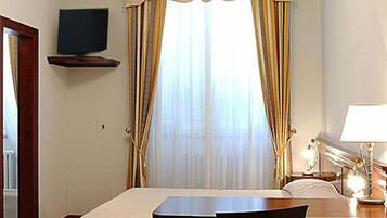 A hotel room with a bed, a desk, a chair, a TV mounted on a shelf, and a window with curtains.