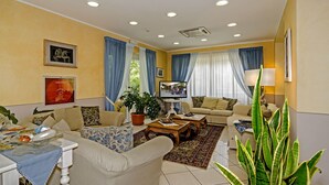 A well-lit living room with a sofa, armchairs, a coffee table, and a TV. There are curtains, a potted plant, and a rug. - Hotel Nautilus (Forte dei Marmi)