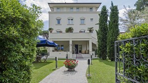 A two-story building with a covered entrance, a paved pathway leading to the entrance, and a flower bed with red flowers. - Hotel Nautilus (Forte dei Marmi)