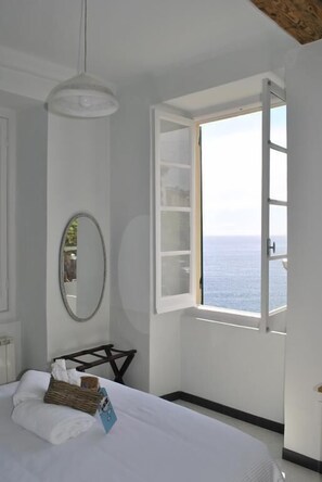 A bedroom with a bed, a bedside table, a mirror, and a window with a view of the sea.