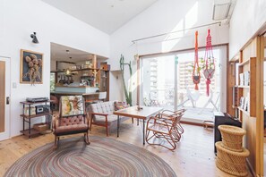 A spacious living area with a wooden dining table, comfortable seating, and a large window offering a view of the city. - SMALL TOWN HOTEL Hakodate (Hakodate)