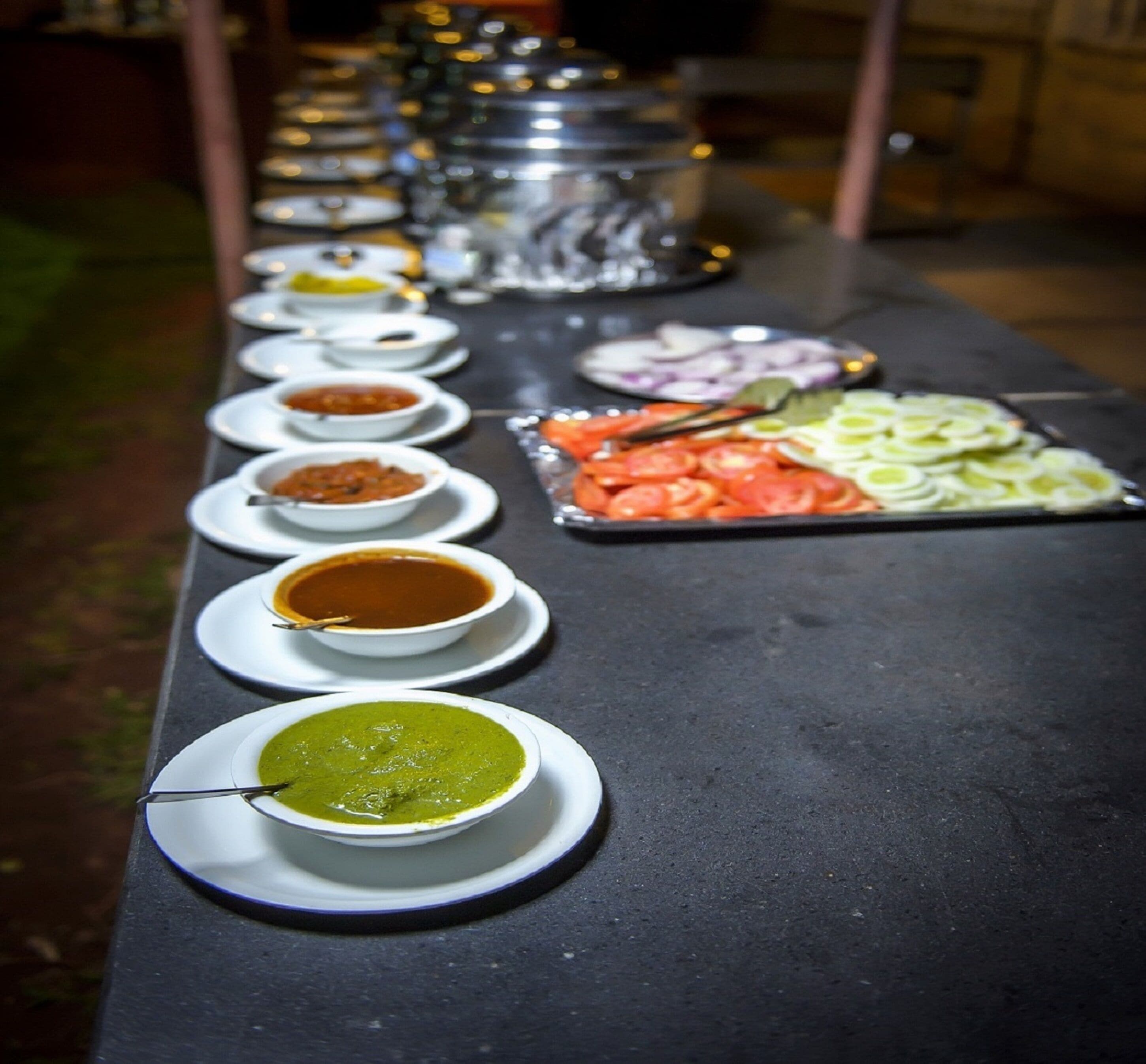 Una mesa con diferentes platos y condimentos, entre ellos una sopa verde, tomates en rodajas y cebollas.
