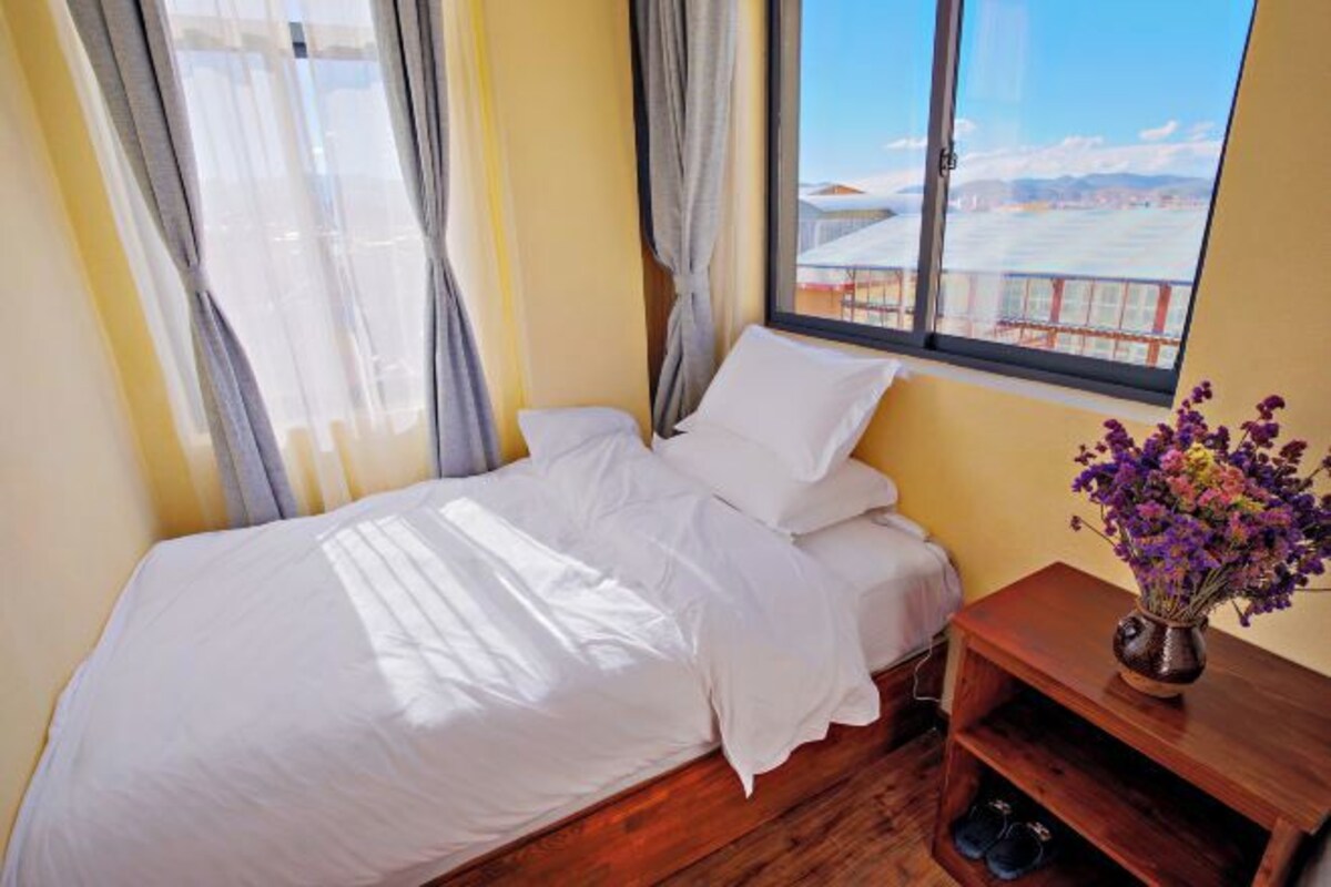 Ein Schlafzimmer mit Bett, einem Holfnachtkästchen, einer Blumenvase und einem Fenster mit Blick auf ein Gebäude und Berge.
