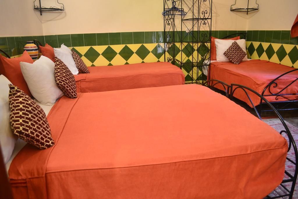 Una stanza con due letti, ognuno con una coperta arancione e cuscini decorati. Si nota una sedia in metallo e un tavolino con un vaso. Le pareti presentano un motivo a scacchi verde e giallo.