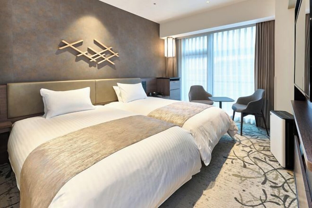 A hotel room with two beds, a desk, a chair, and a large window.