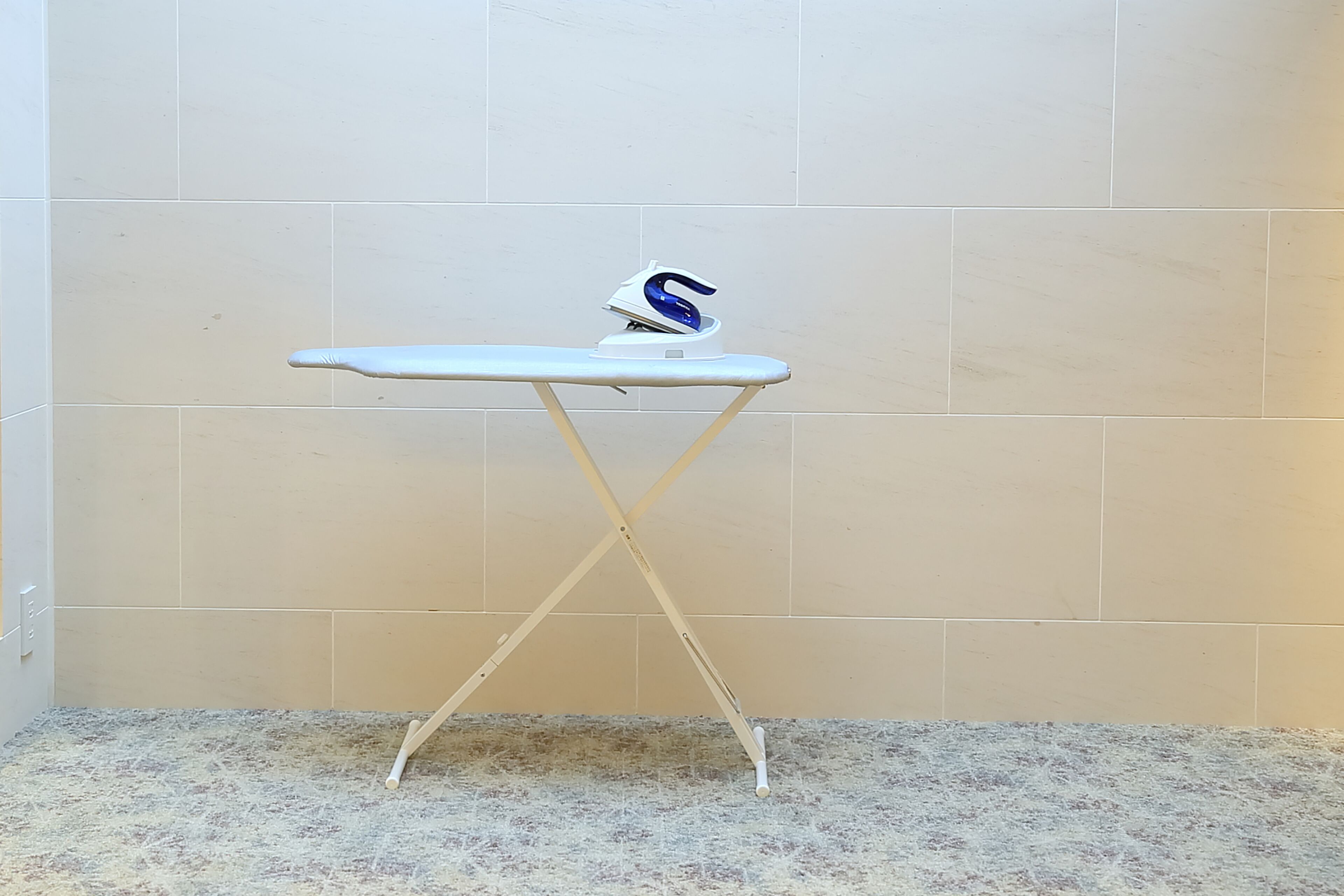 A white ironing board with a blue and white iron on top, placed against a tiled wall.