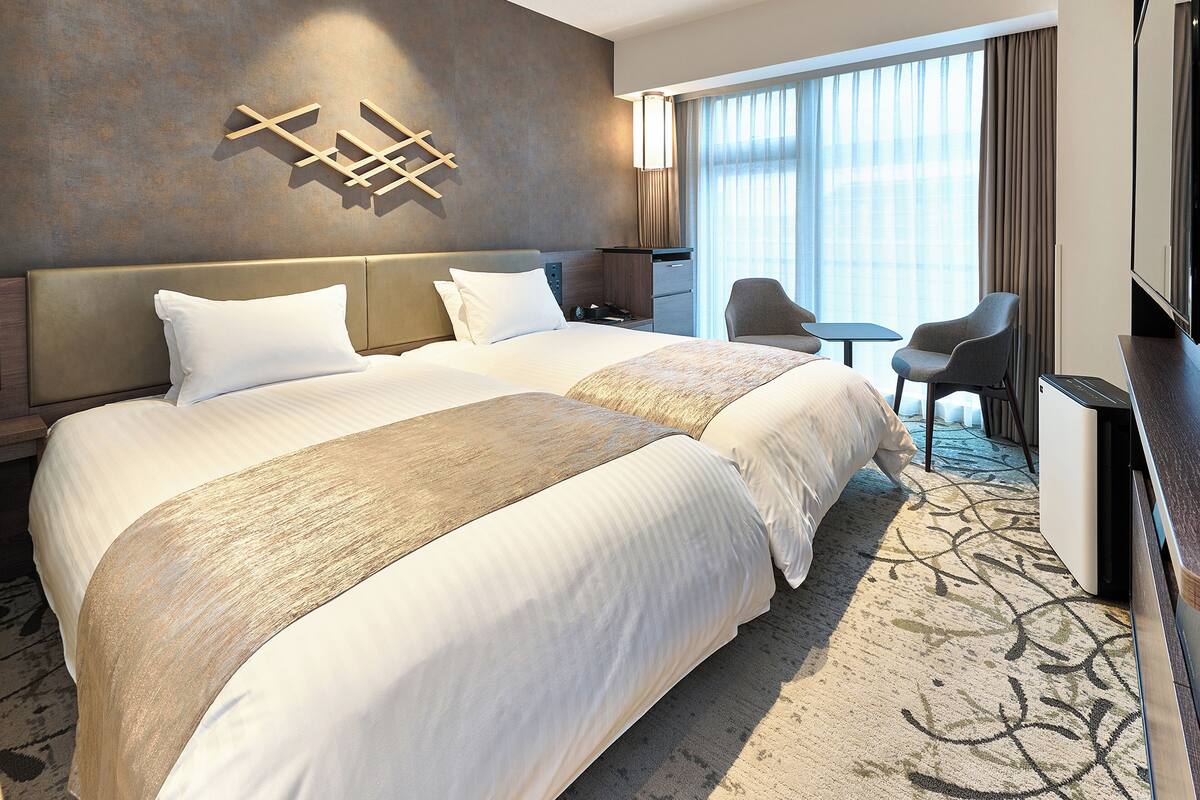 A hotel room with two beds, a desk, a chair, and a large window with curtains.