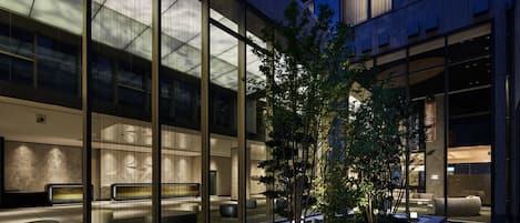 A modern building with large glass windows, a landscaped garden with trees and white pebbles, and outdoor seating areas.
