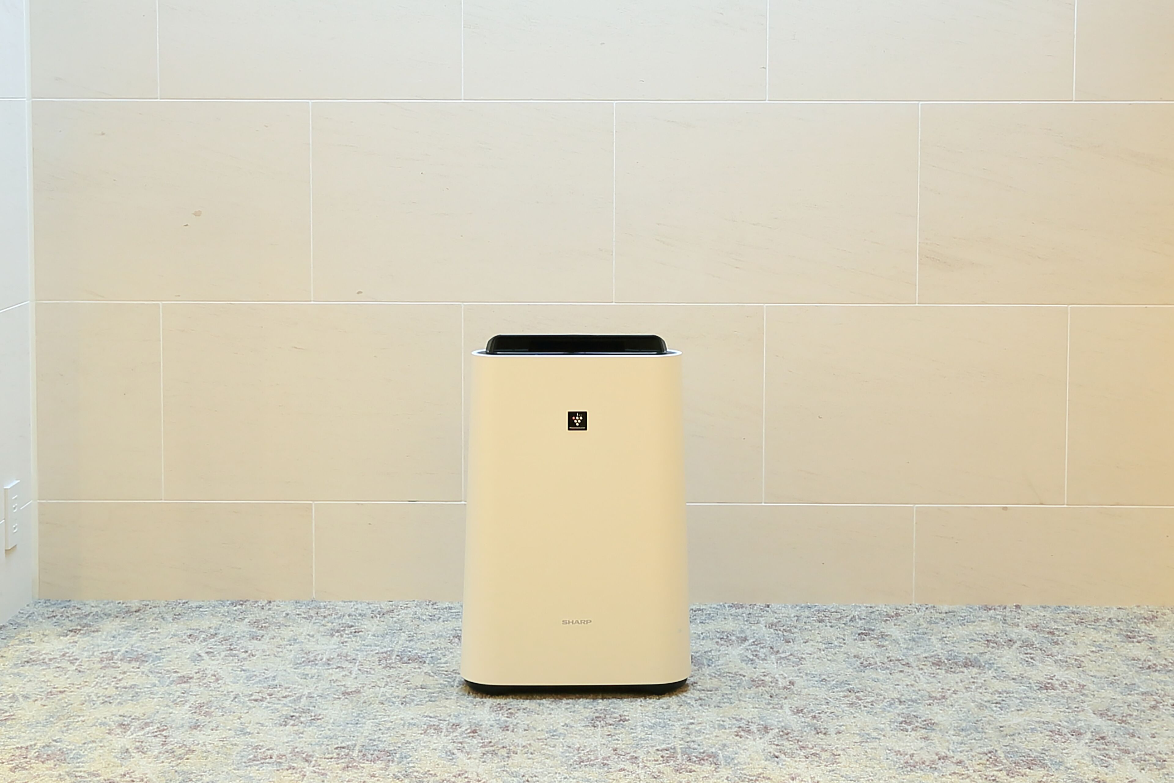 A compact, beige air purifier with a control panel on top, placed on a speckled countertop against a plain tiled wall.
