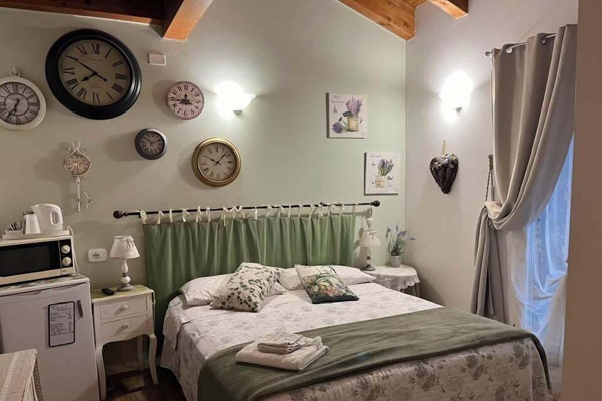 A bedroom with a bed, a nightstand, a dresser, a microwave, and multiple clocks on the wall.