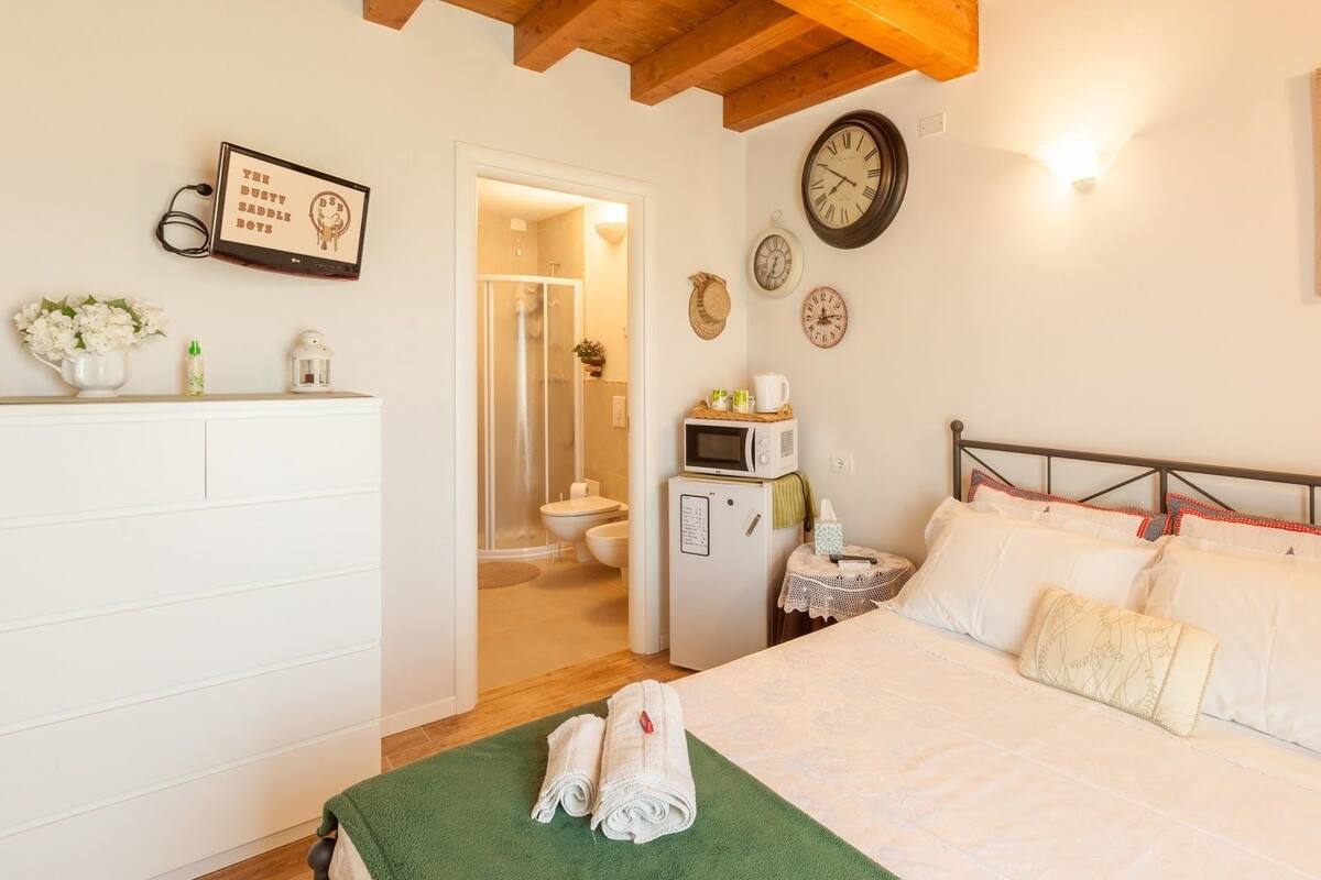 A bedroom with a bed, a bedside table, a microwave, a clock, and a door leading to a bathroom.