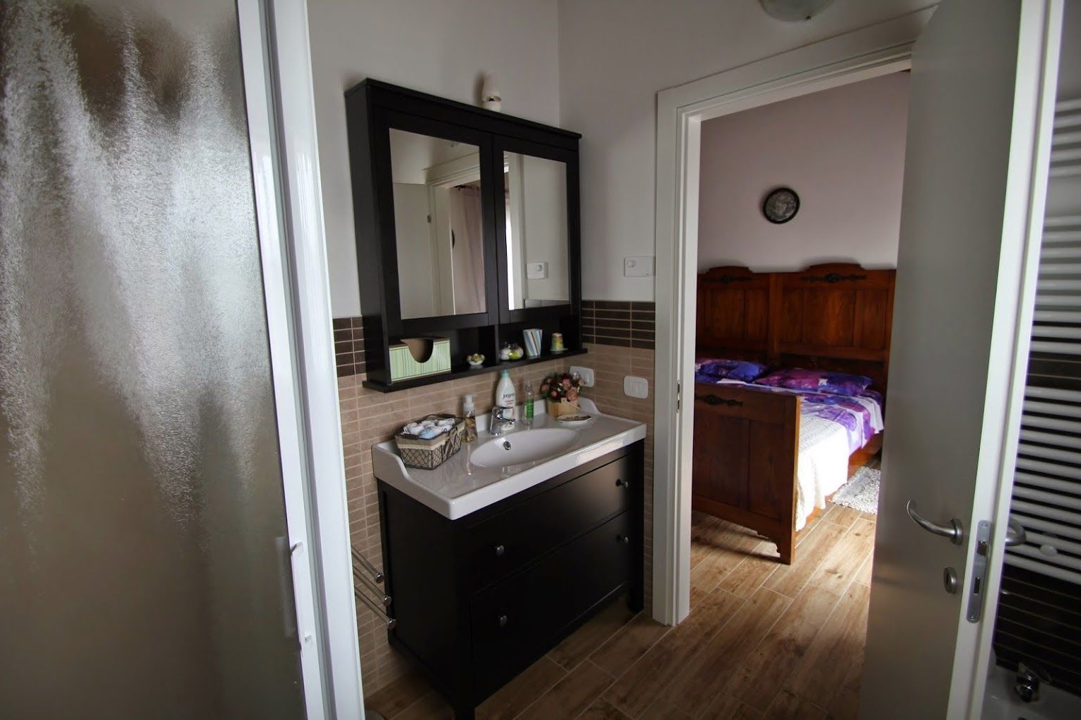 A bathroom with a double sink, a mirror, and a wooden door leading to a bedroom with a bed and a wooden headboard.