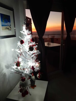 Ein weißer Weihnachtsbaum mit roten Verzierungen und Lichtern, platziert in einem Raum mit Blick auf einen Sonnenuntergang durch ein Fenster.