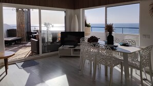 Ein modernes Wohnzimmer mit Fernseher, weißen Möbeln und großen Fenstern mit Meerblick.