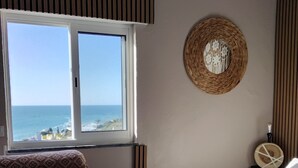 Ein Schlafzimmer mit einem großen Fenster, das Meerblick bietet. Es gibt ein Bett mit weißer Bettdecke, einen Nachttisch mit einer Lampe und eine gewebte Wanddekoration.