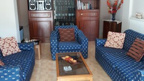 A living room with two blue sofas, a glass coffee table, wooden cabinets, and a bookshelf. - Villa near Athens Airport & Rafina (Spata-Artemida)
