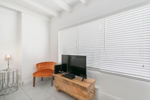 Una sala moderna con un televisor de pantalla plana grande sobre un mueble de madera, una mesita redonda con una lámpara y un sillón naranja cómodo.
