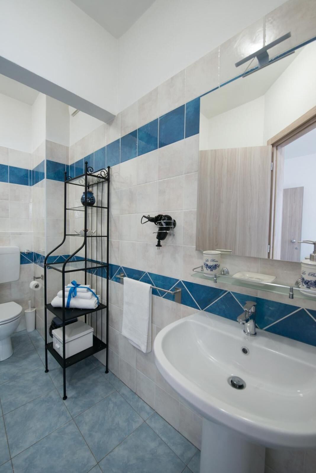 A bathroom with a white sink, a black metal shelf with towels, a toilet, and a mirror with a wooden frame.