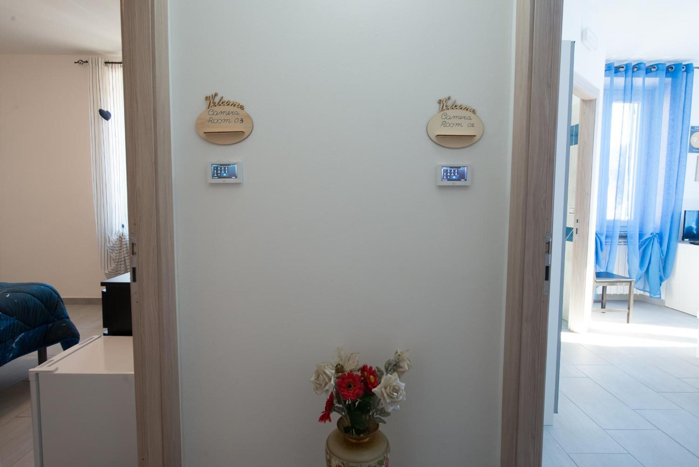 Un área de recepción con dos letreros que indican 'Welcome Camera Room 03' y 'Welcome Camera Room 04'. En primer plano, una pequeña mesa con un jarrón de flores.