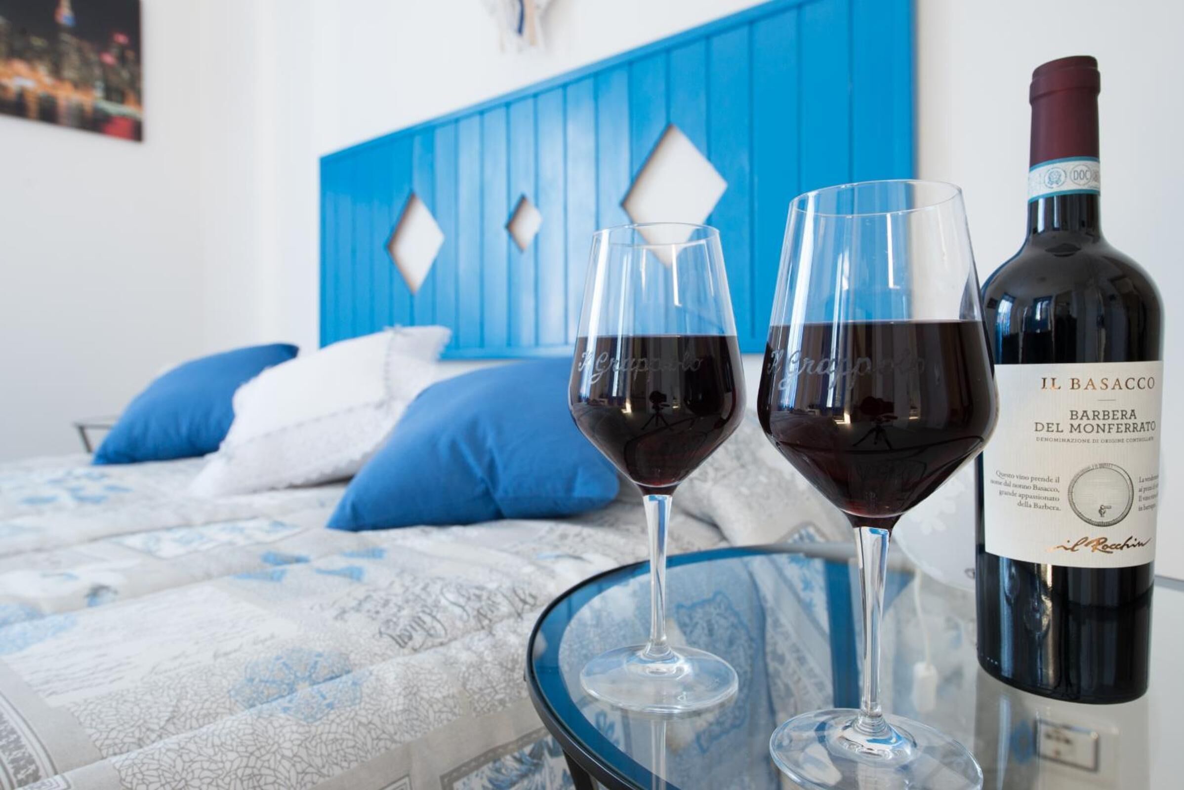Uma cama com cabeceira azul, dois copos de vinho sobre uma mesa de vidro e uma garrafa de vinho.