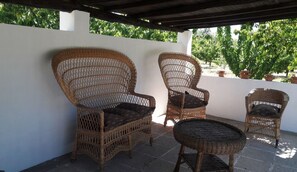 Wicker chairs and a small table on a patio with a white wall and potted plants in the background.